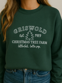 A woman wearing a green sweatshirt with white text that says "GRISWOLD est. 1989 CHRISTMAS TREE FARM little full, lotta lap."