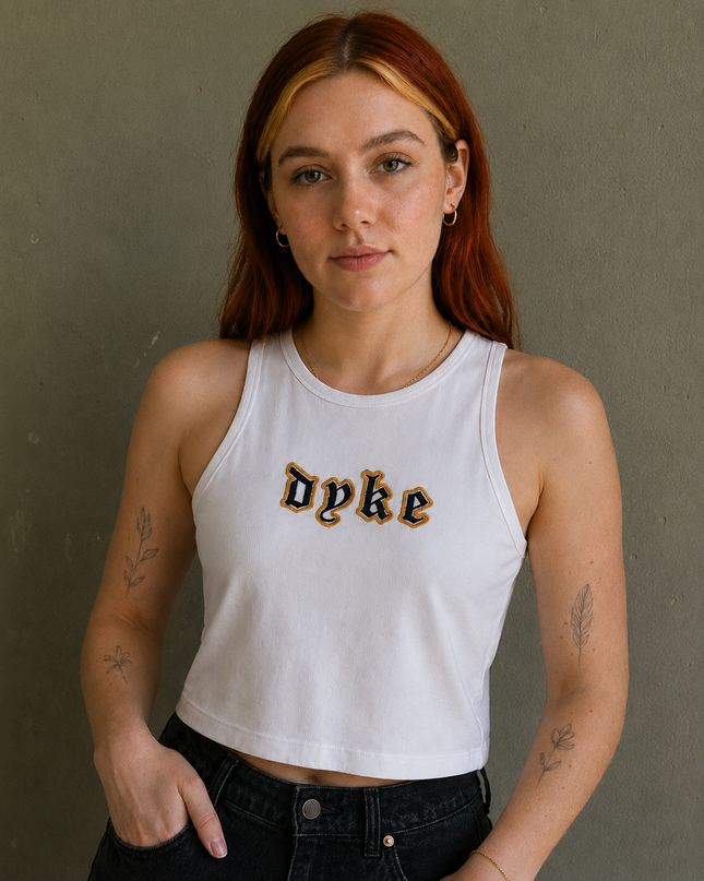 A young woman with red hair wearing a white tank top with the word "Dyke" printed on it, posing against a gray wall.
