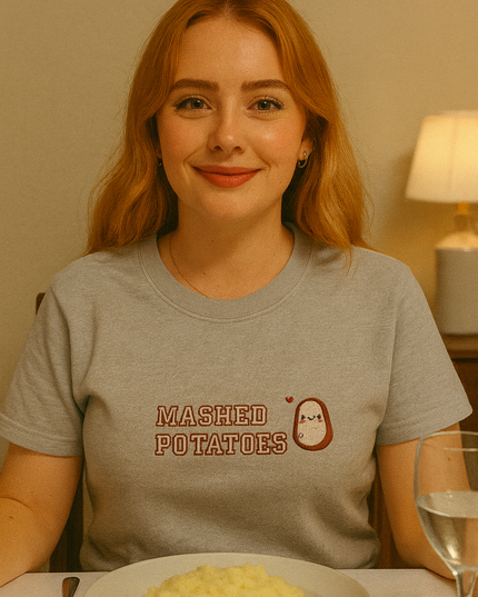 A woman with red hair is sitting at a table with a plate of mashed potatoes in front of her. She is wearing a gray t-shirt with the text "Mashed Potatoes" printed on it.