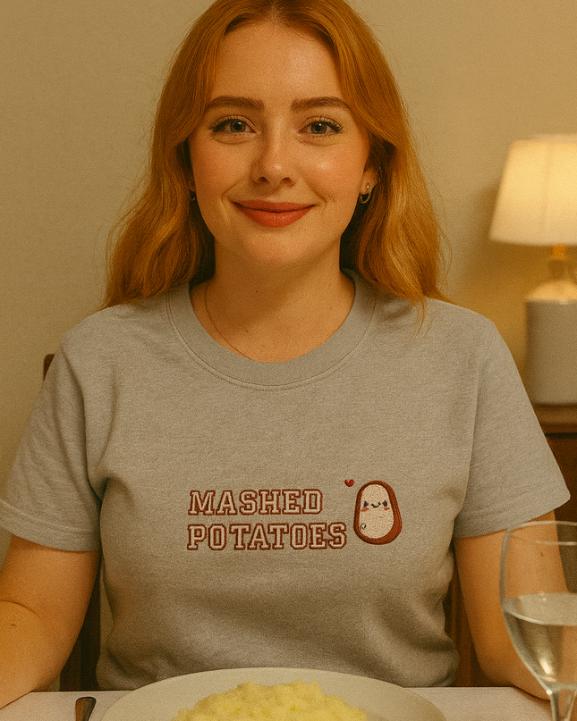 A woman with red hair is sitting at a table with a plate of mashed potatoes in front of her. She is wearing a gray t-shirt with the text "Mashed Potatoes" printed on it.