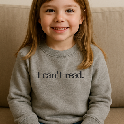 A young girl with long brown hair is sitting on a couch, smiling and wearing a gray sweatshirt with the text "I can't read" printed on it.