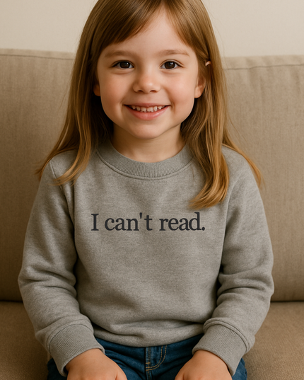 A young girl with long brown hair is sitting on a couch, smiling and wearing a gray sweatshirt with the text "I can't read" printed on it.