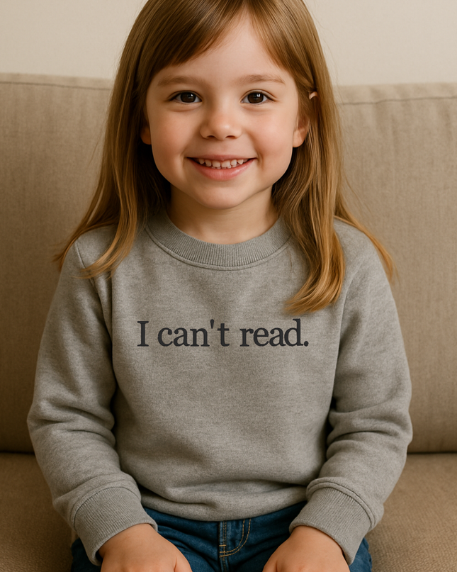 A young girl with long brown hair is sitting on a couch, smiling and wearing a gray sweatshirt with the text "I can't read" printed on it.