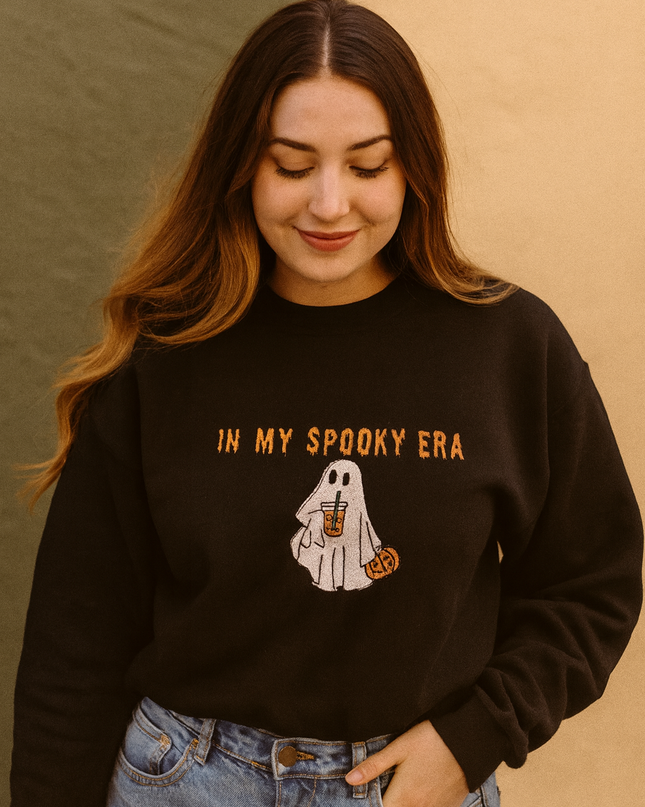 A woman wearing a black sweatshirt with a graphic of a ghost and the text "In my spooky era" is smiling and looking down.