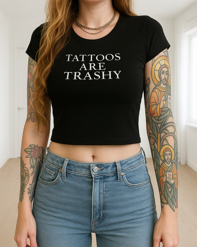 A woman wearing a black crop top with the text "Tattoos are Trashy" printed on it, standing in a room with her arms crossed.