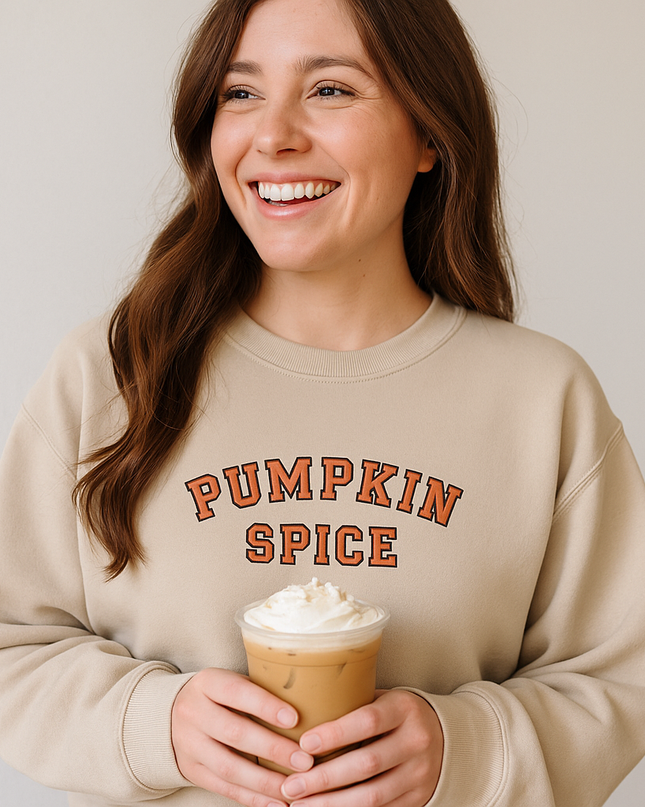 A woman with long brown hair is smiling and holding a cup of iced coffee with whipped cream on top. She is wearing a beige sweater with the text "PUMPKIN SPICE" printed on it.