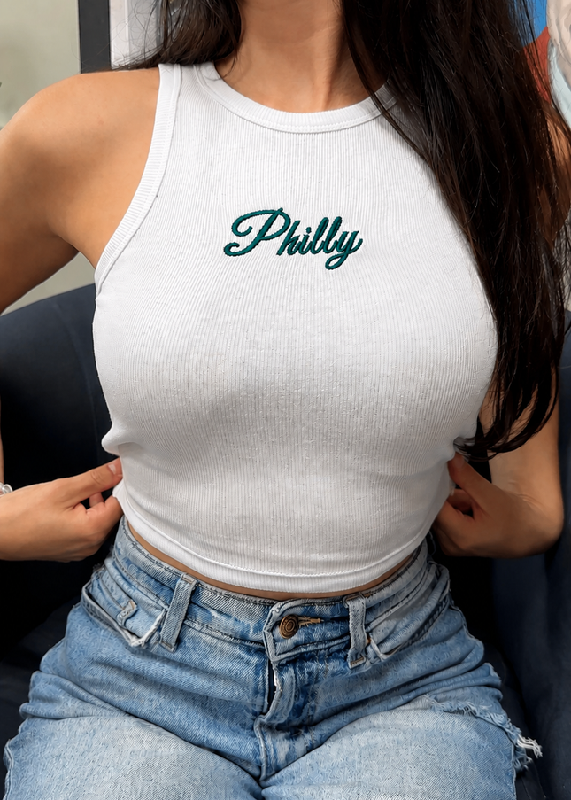 A woman wearing a white tank top with the word "Philly" printed on it, paired with blue jeans.