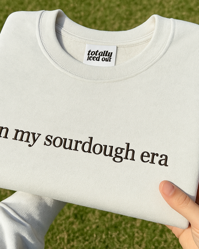 A person's hand holding a white t-shirt with the text "in my sourdough era" printed on it.