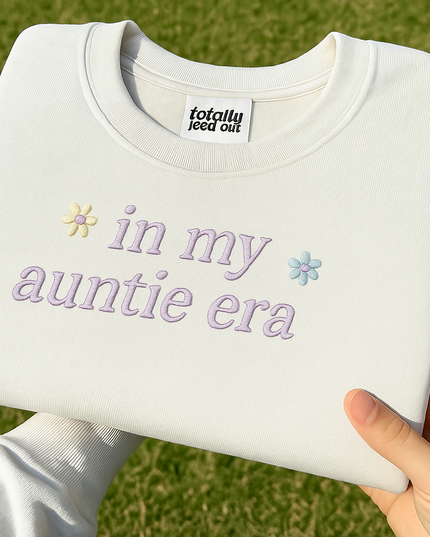 A person's hand holding a white t-shirt with the text "in my auntie era" embroidered on it.