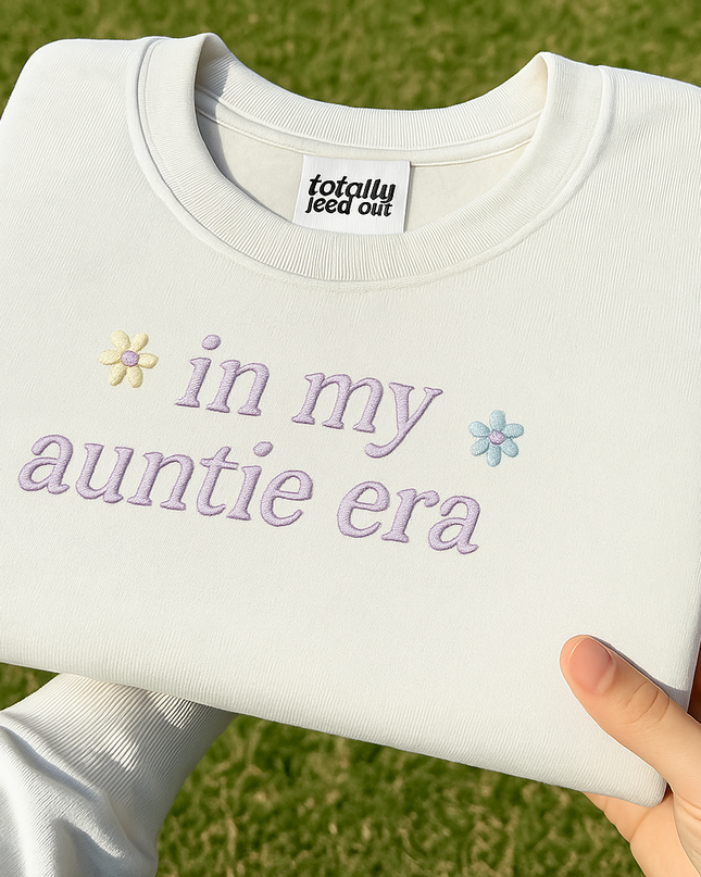 A person's hand holding a white t-shirt with the text "in my auntie era" embroidered on it.