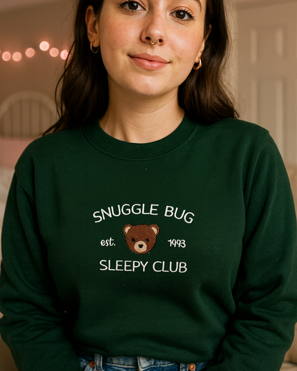 A young woman wearing a green sweatshirt with the text "SNuggle Bug" and "Est. 1993 SLEEPY CLUB" printed on it.