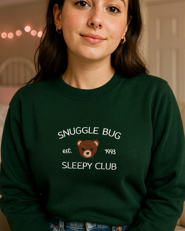 A young woman wearing a green sweatshirt with the text "SNuggle Bug" and "Est. 1993 SLEEPY CLUB" printed on it.