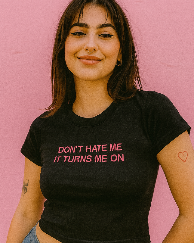 A young woman with long brown hair is smiling and wearing a black t-shirt with the text "Don't Hate Me It Turns Me On" printed on it.