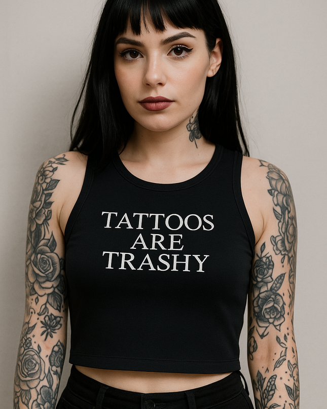 A young woman with long dark hair and tattoos on her arms is wearing a black crop top with the text "Tattoos are Trashy" printed on it.