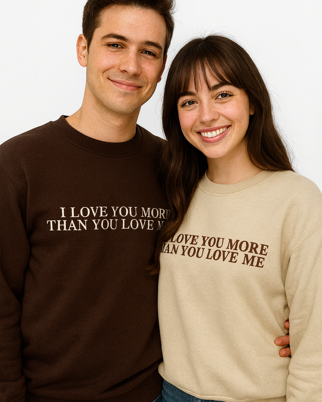 A man and a woman standing next to each other, both smiling and wearing matching sweaters with the text "I LOVE YOU MORE THAN YOU LOVE ME" printed on them.