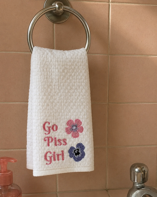 A white hand towel with the text "Go Piss Girl" embroidered on it, hanging on a towel rack next to a pink soap dispenser.