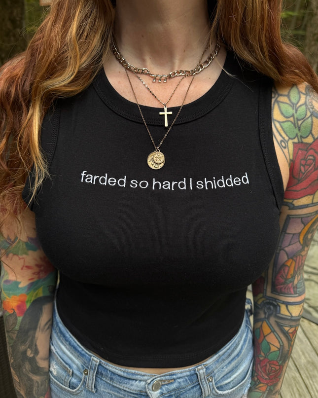 A person with long, brown hair wearing a black tank top with the text "farded so hard I shidered" printed on it. They are also wearing a cross necklace and have tattoos on their arms.