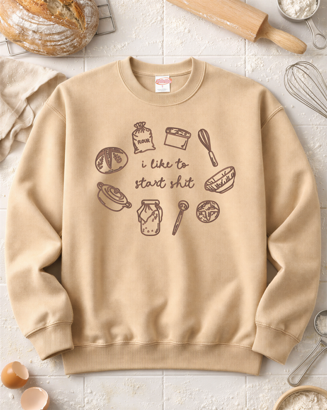 a beige sweatshirt with a humorous text printed on it, surrounded by various baking-related items such as a rolling pin, a whisk, and a bowl of flour.