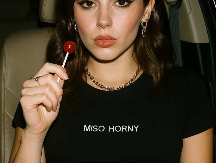 A young woman with long brown hair is sitting in a car, holding a red lollipop. She is wearing a black t-shirt with the text "MISO HORNY" printed on it.