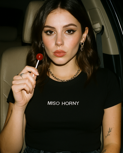 A young woman with long brown hair is sitting in a car, holding a red lollipop. She is wearing a black t-shirt with the text "MISO HORNY" printed on it.
