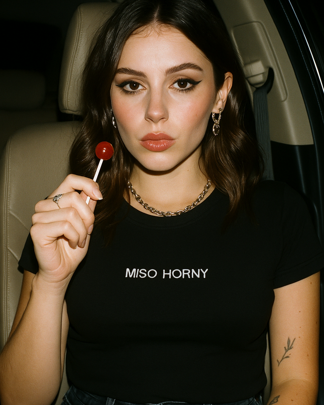 A young woman with long brown hair is sitting in a car, holding a red lollipop. She is wearing a black t-shirt with the text "MISO HORNY" printed on it.