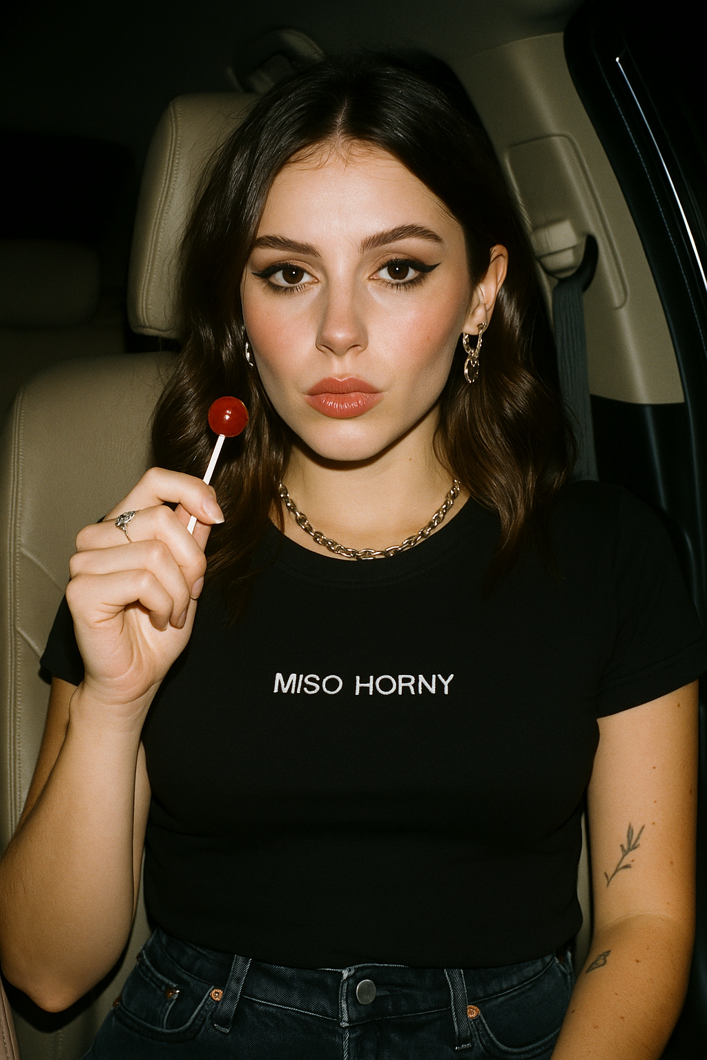 A young woman with long brown hair is sitting in a car, holding a red lollipop. She is wearing a black t-shirt with the text "MISO HORNY" printed on it.