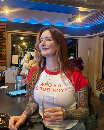 A woman with long brown hair wearing a red and white t-shirt with the text "Who's a good boy?" is sitting at a table in a bar, holding a glass of beer.