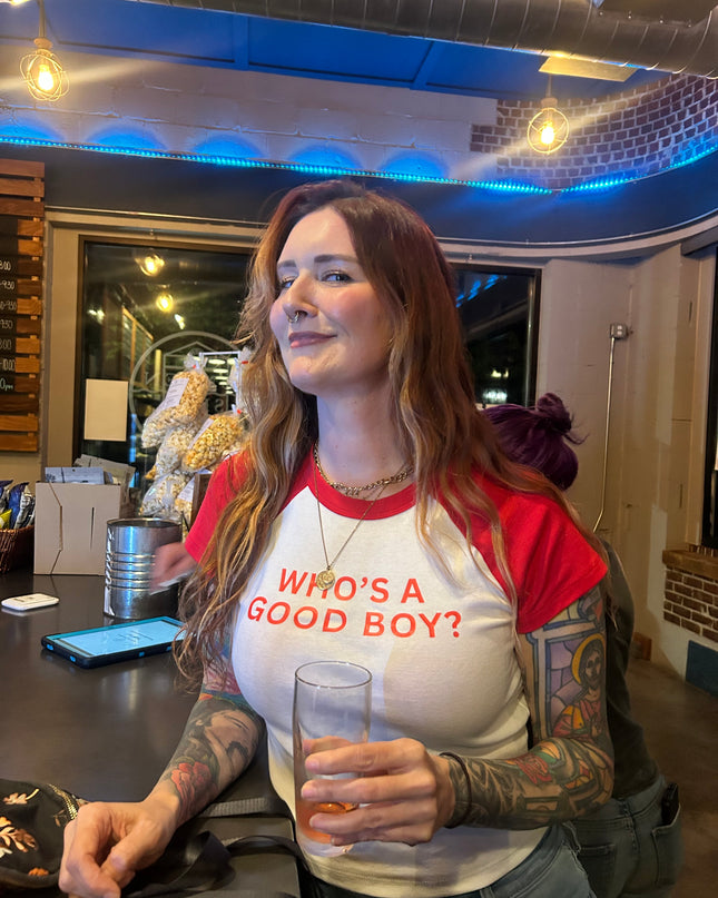 A woman with long brown hair wearing a red and white t-shirt with the text "Who's a good boy?" is sitting at a table in a bar, holding a glass of beer.