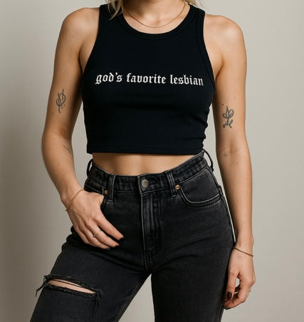 A woman wearing a black crop top with the text "God's favorite Lesbian" printed on it, paired with distressed black jeans.