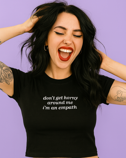 A woman with long dark hair is smiling and making a funny face, wearing a black crop top with white text.
