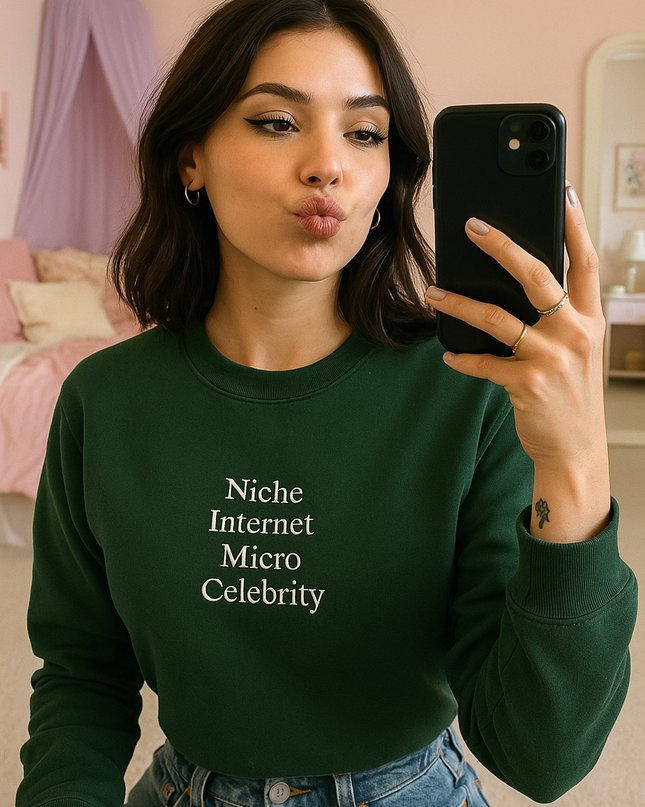 A young woman in a green sweater and denim shorts is taking a selfie with a black smartphone, while a pink canopy can be seen in the background.