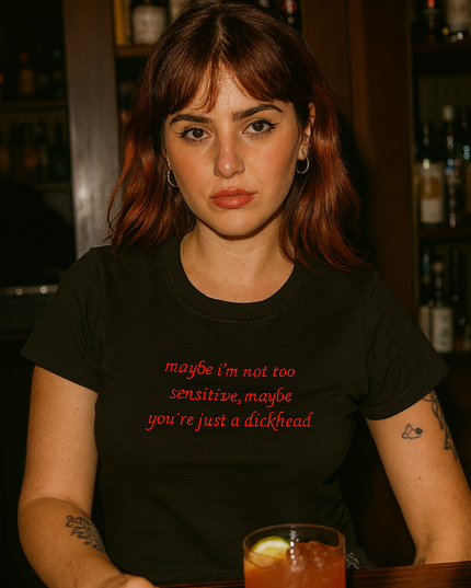 A woman with long brown hair wearing a black t-shirt with text on it, sitting at a bar with a glass of drink in front of her.