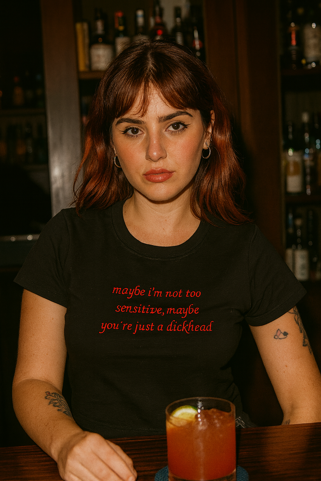 A woman with long brown hair wearing a black t-shirt with text on it, sitting at a bar with a glass of drink in front of her.