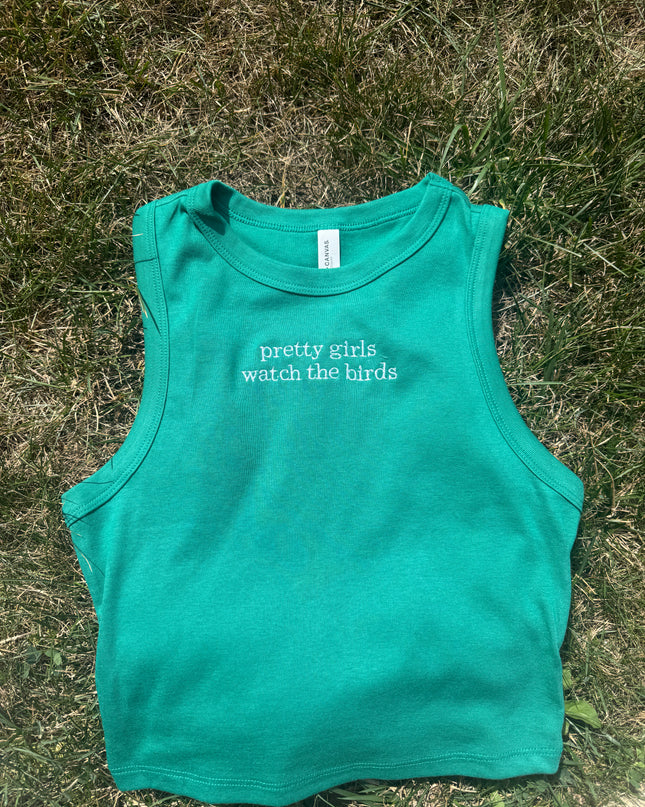 A green tank top with the text "prettily girls watch the birds" printed on it, lying on the grass.