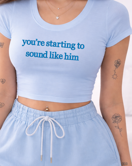 A woman wearing a light blue crop top with the text "you're starting to sound like him" printed on it, paired with a pair of blue shorts. She has tattoos on her arms and a necklace around her neck.