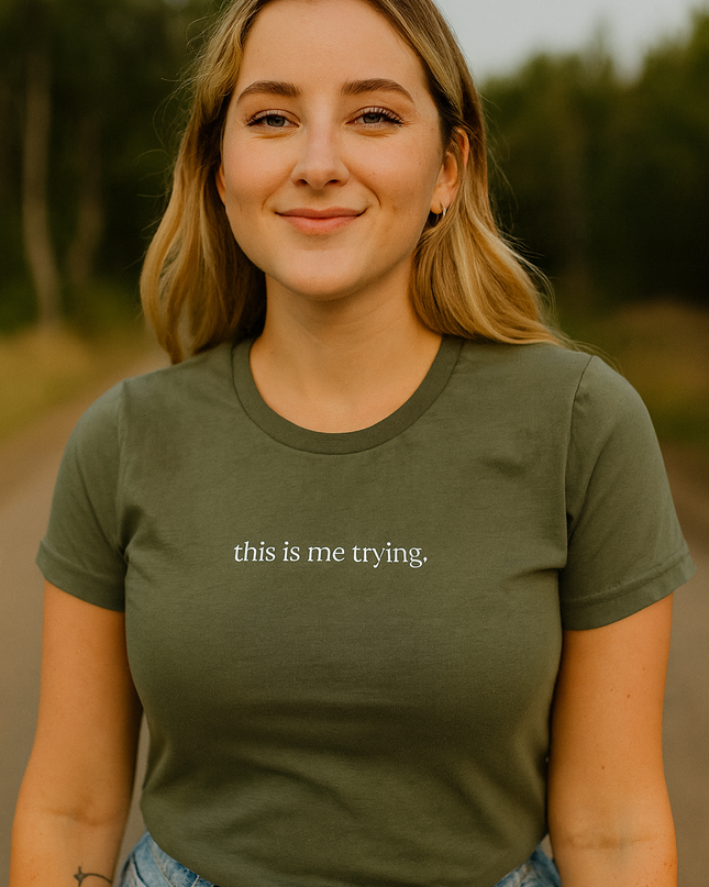 A young woman with blonde hair is smiling and wearing a green t-shirt with the text "this is me trying" printed on it.