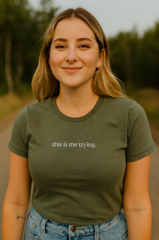 A young woman with blonde hair is smiling and wearing a green t-shirt with the text "this is me trying" printed on it.