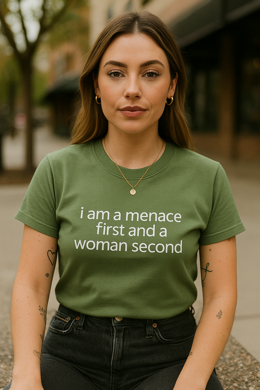 A woman wearing a green t-shirt with the text "I am a manace first and a woman second" stands in front of a building.
