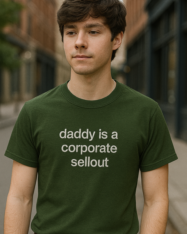 A young man wearing a green t-shirt with the text "daddy is a corporate sellout" stands in an urban setting.