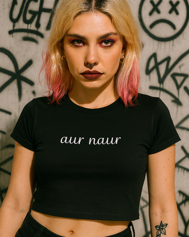 A young woman with blonde hair and makeup stands in front of a graffiti-covered wall, wearing a black crop top with the text "aur naur" printed on it.