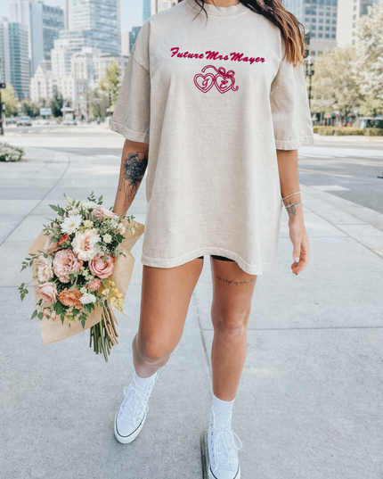 A woman wearing a beige t-shirt with the text "Future Mrs. Mayer" and holding a bouquet of flowers is walking on a sidewalk in an urban setting.