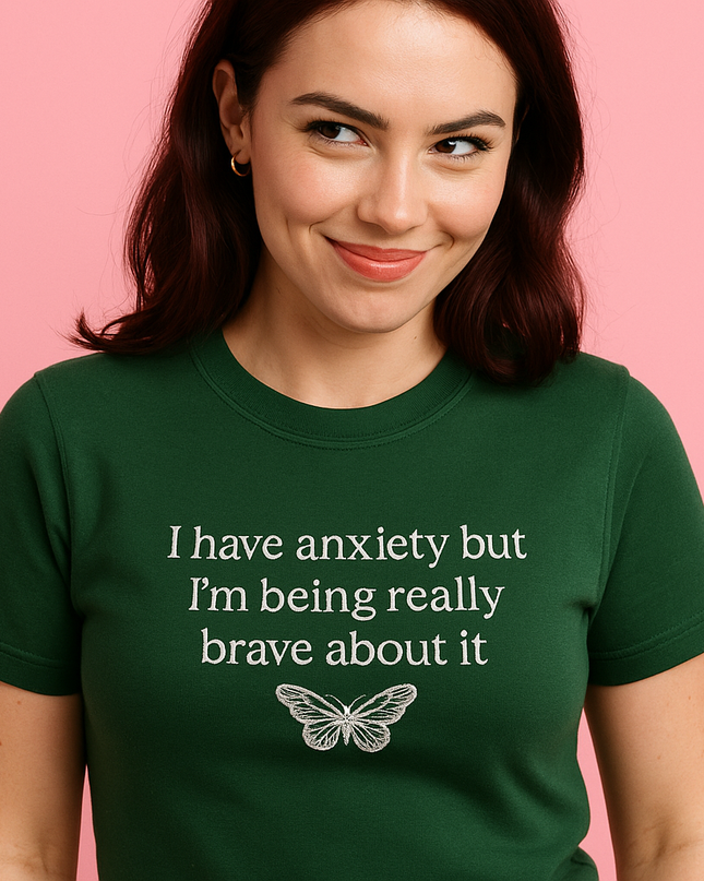 A woman wearing a green t-shirt with the text "I have anxiety but I'm being really brave about it" printed on it, smiling at the camera.