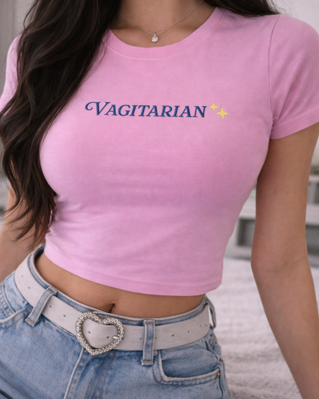 A woman wearing a pink crop top with the word "Vagitarian" printed on it, paired with denim shorts and a heart-shaped belt buckle.