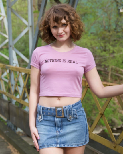 A young woman wearing a pink crop top with the text "Nothing is real" and a denim mini skirt, standing on a bridge with a metal railing in the background.