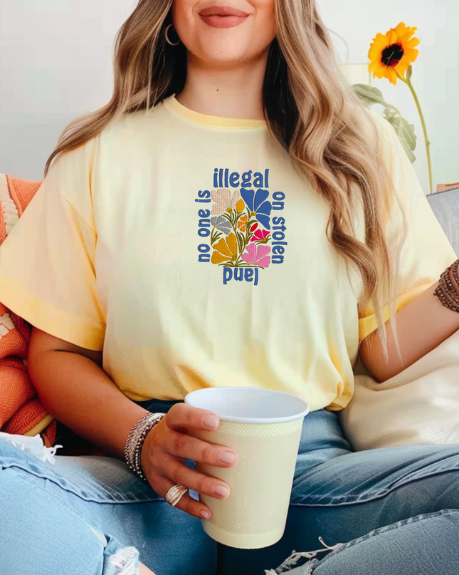 A woman wearing a yellow t-shirt with a graphic design and holding a white cup, sitting on a couch with a yellow flower in the background.