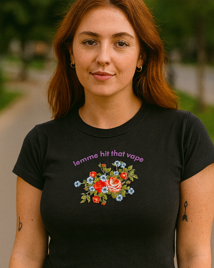 A young woman with long red hair wearing a black t-shirt with a floral design and the text "lemme hit that vapor" stands outdoors.