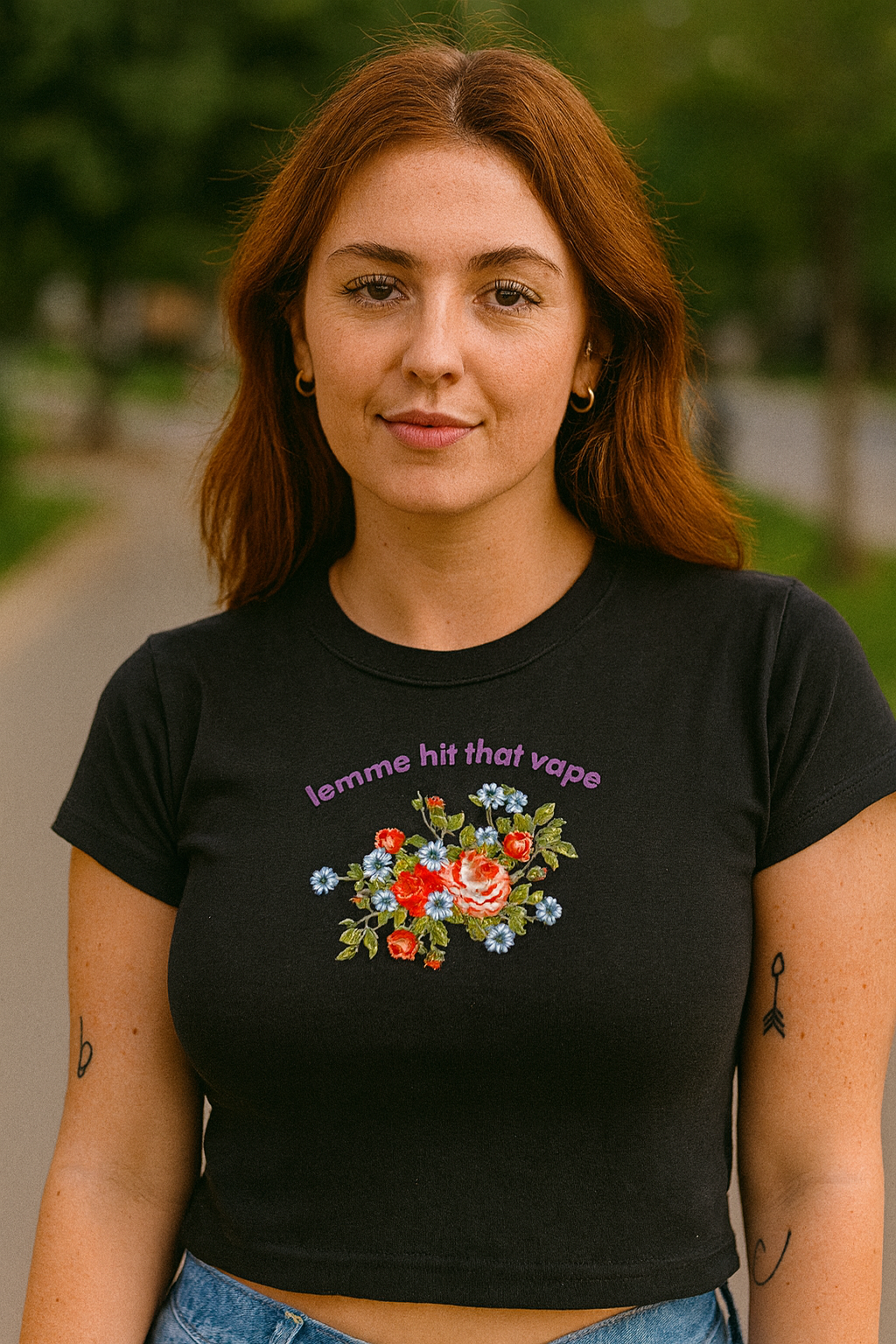 A young woman with long red hair wearing a black t-shirt with a floral design and the text "lemme hit that vapor" stands outdoors.