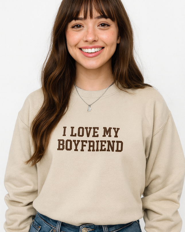 A smiling woman with long brown hair wearing a beige sweatshirt with the text "I LOVE MY BOYFRIEND" printed on it.