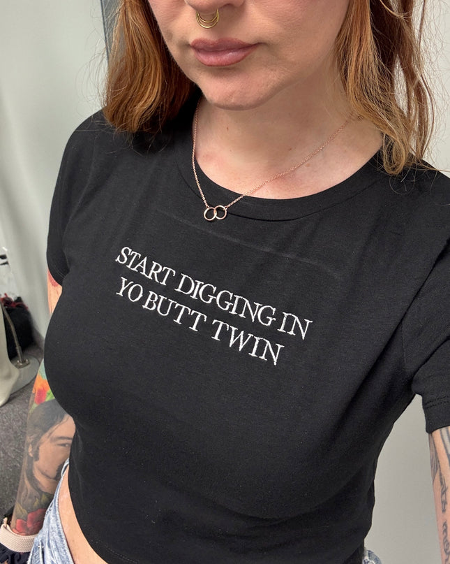 A woman wearing a black t-shirt with the text "START DIGGING IN YO BUTT TWIN" printed on it.
