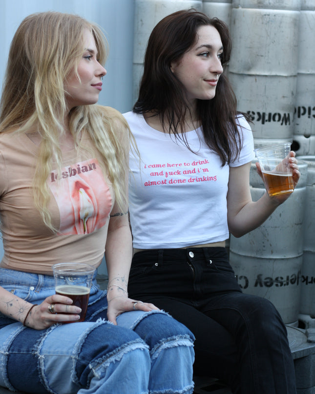 Two young women are sitting together, one holding a drink, in front of a white wall with text on it.